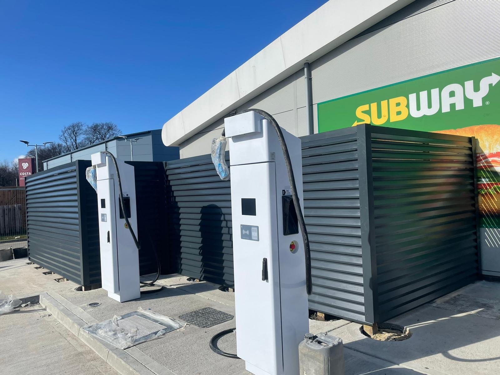 Electric vehicle charging stations are lined up outside a building with a Subway logo. Positioned on a concrete surface, they feature modern gray panels—an installation showcasing the work of a commercial fencing contractor.