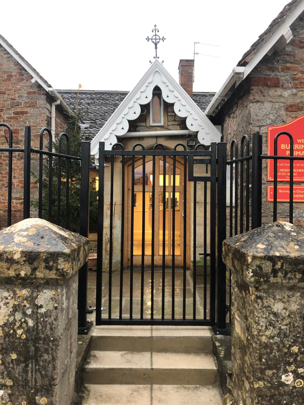 A gated entrance to a small stone church with Gothic-style architecture, featuring a pointed arch doorway and decorative trim. The black metal gate, showcasing expert fencing installation, is flanked by two stone pillars. Inside, warm light emanates from the interior.