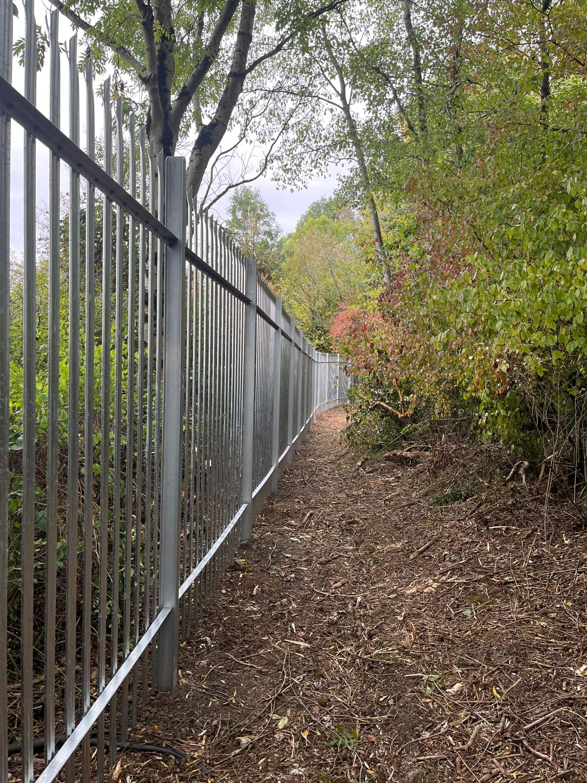 A long, tall metal fence installed by a commercial fencing contractor runs alongside a dirt pathway surrounded by trees and bushes. The foliage is mostly green, with some red and yellow leaves, indicating early autumn. The sky is overcast, giving a muted tone to the scene.