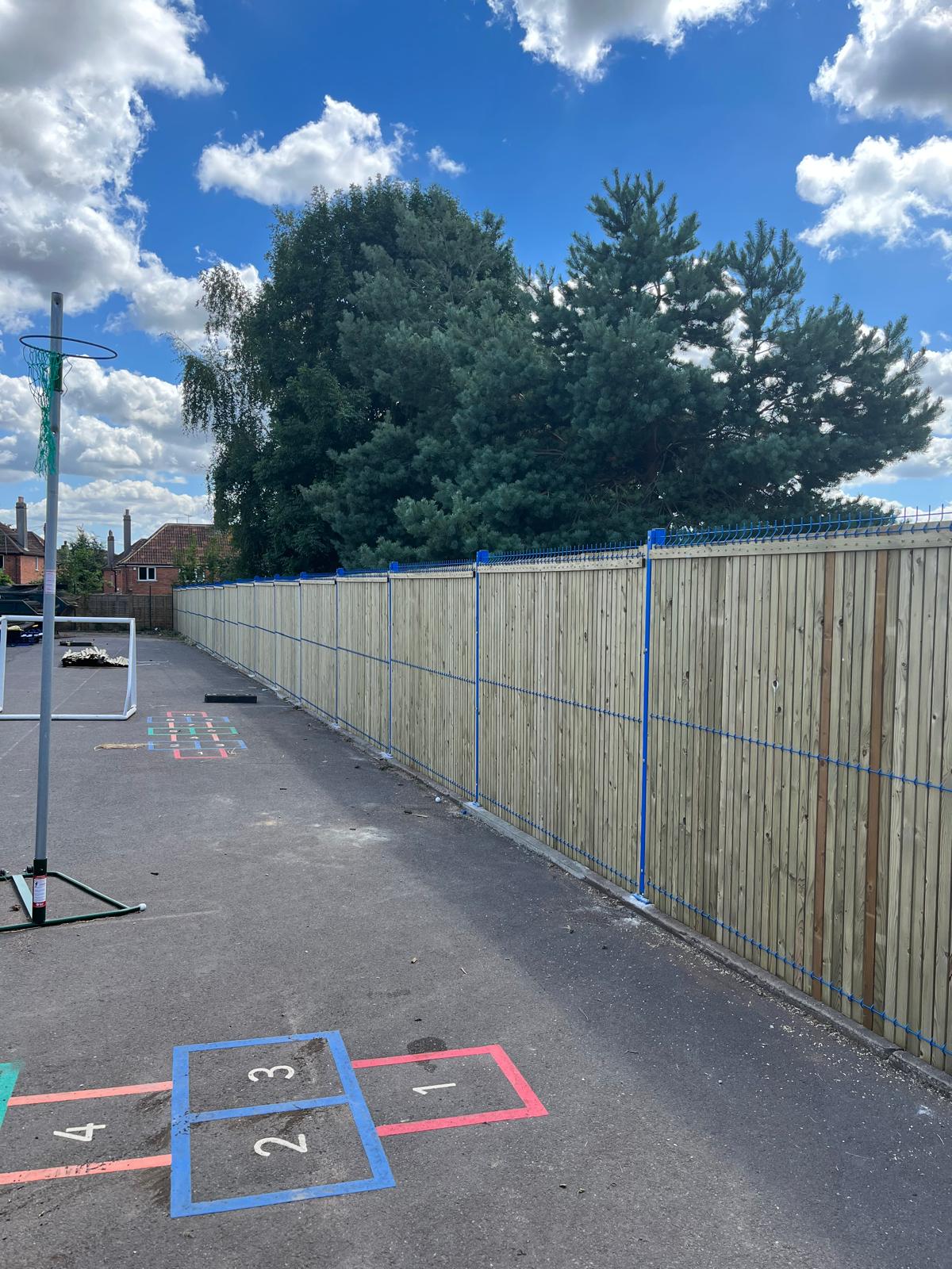 A school playground with hopscotch markings is shown. A basketball hoop is on the left. A tall wooden fence, crafted by a commercial fencing contractor, runs alongside, with trees visible behind it. The sky is blue with scattered clouds.