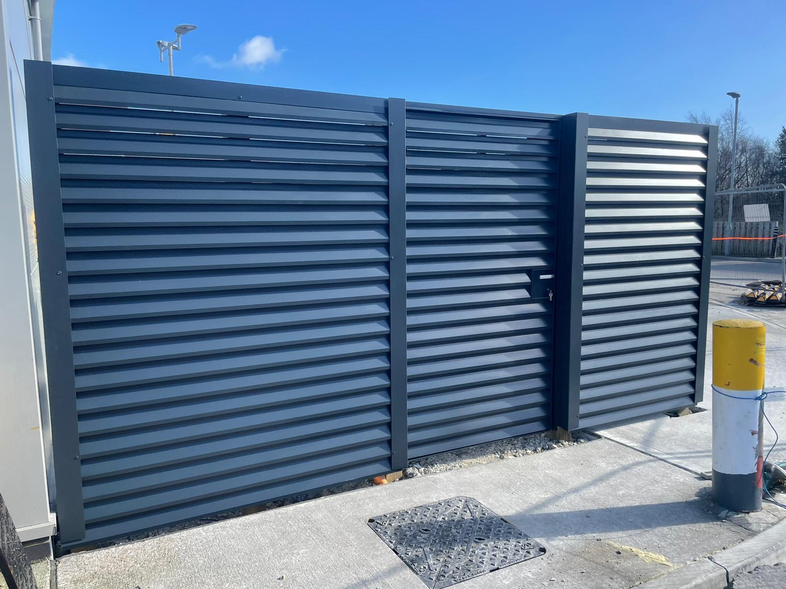 A dark gray louvered gate with three panels stands on a concrete surface, adjacent to a metal post. A drain cover is visible in front. The area showcases new gates as part of a well-planned fencing installation, with trees and a fence under a clear blue sky completing the view.