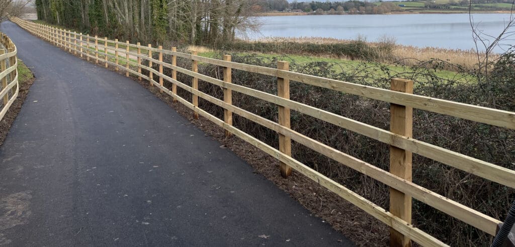 A paved pathway runs beside a lake, bordered by bespoke railings on both sides. The lake is surrounded by grass and trees, with a view of distant hills under a cloudy sky.