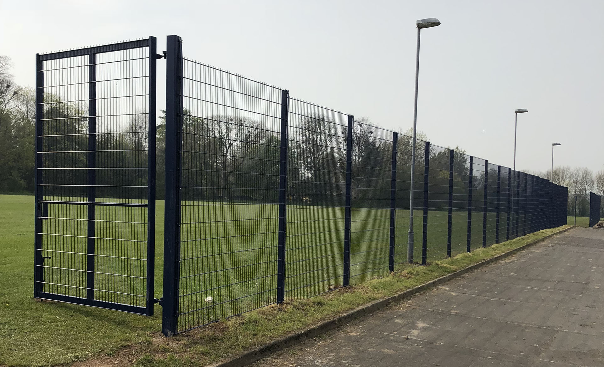 A tall, black metal fence with bespoke railings and a gate separates a grassy field from a paved pathway. The fence runs alongside the path, illuminated by streetlights. Trees are visible in the background under an overcast sky.