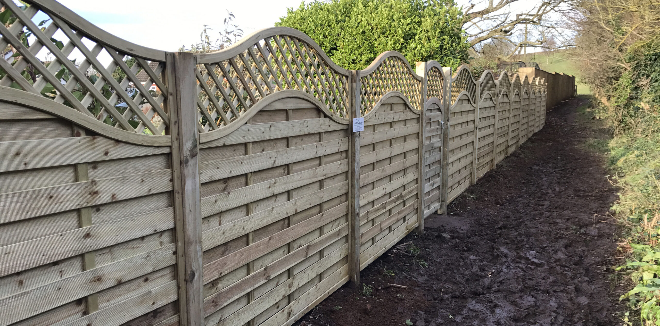 A wooden fence with wavy lattice tops, crafted by a skilled commercial fencing contractor, lines a narrow muddy path. Tall bushes and trees are visible on the opposite side. The fence stretches into the distance under a clear sky.