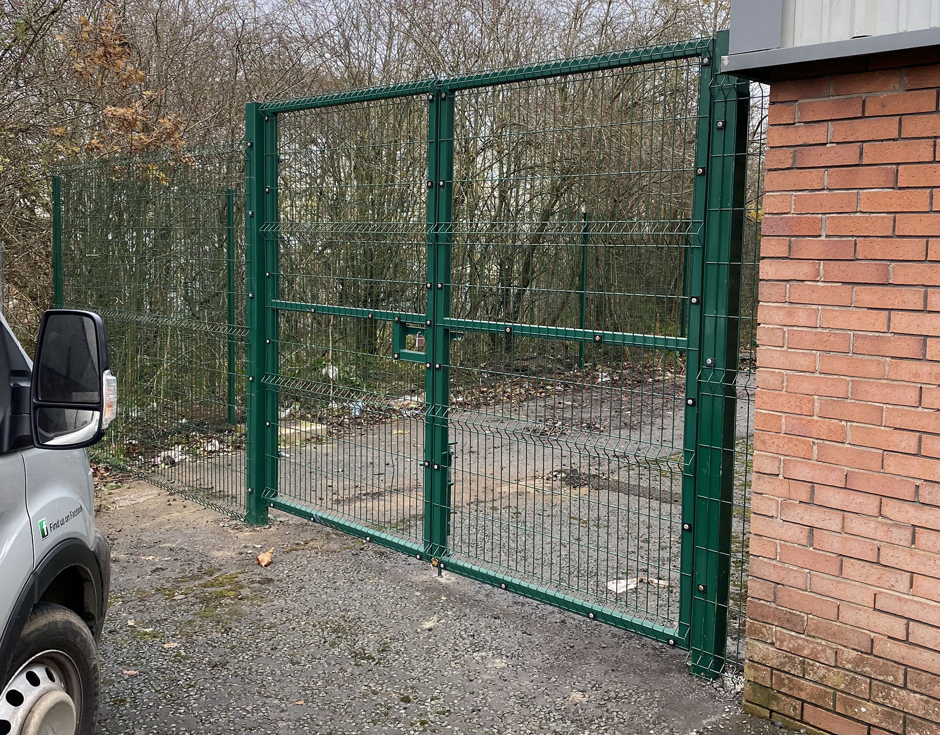 A green metal fence with new gates stands between a brick building and a parking area. The gates, crafted by a commercial fencing contractor, are closed, with a white van partially visible on the left. Leafless trees and scattered litter are in the background.