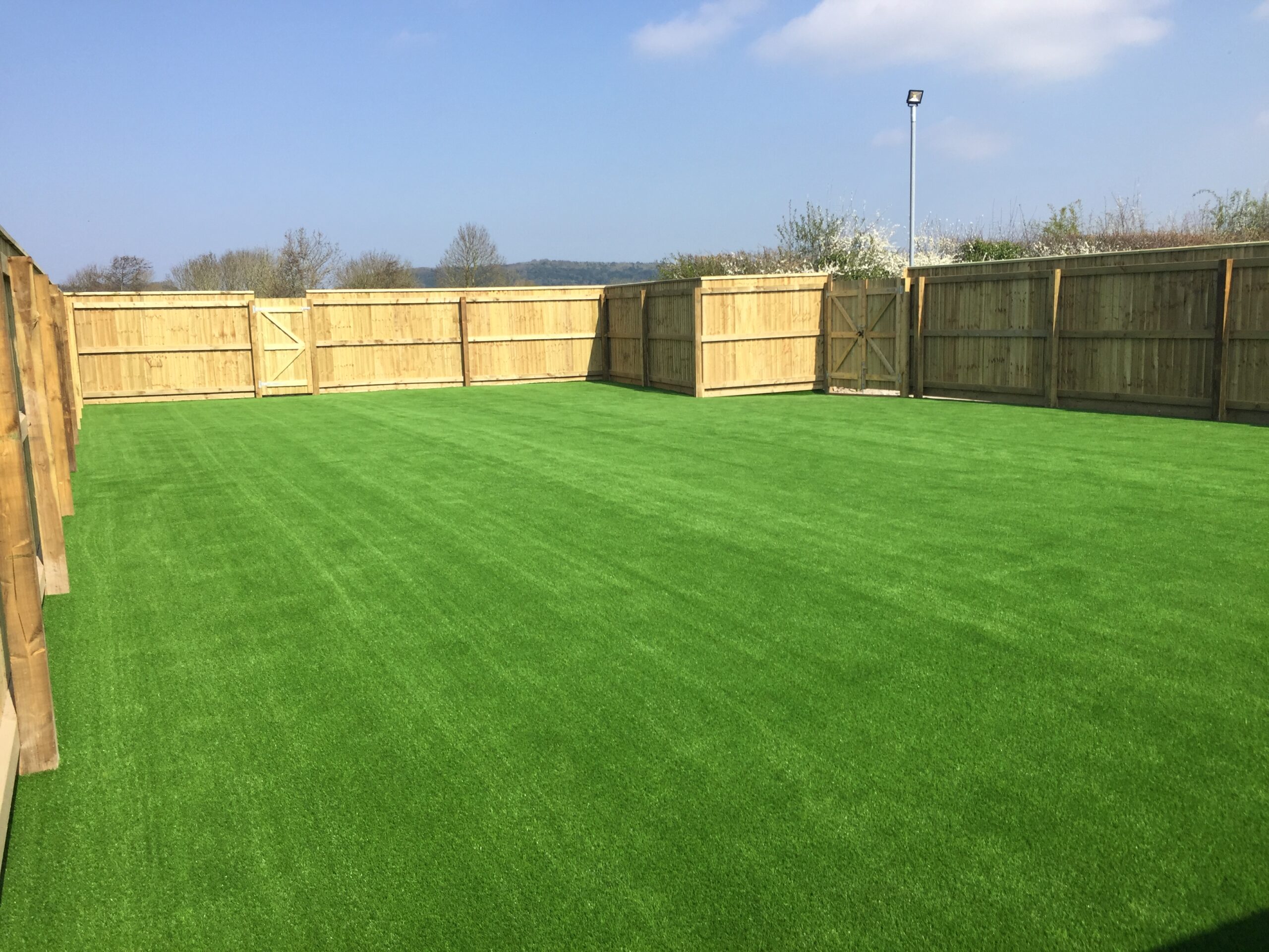 A spacious outdoor area with lush green artificial grass, enclosed by expertly crafted fencing installation. The sky is clear and blue, and the scene is well-lit by sunlight, enhancing the beauty of this serene retreat.