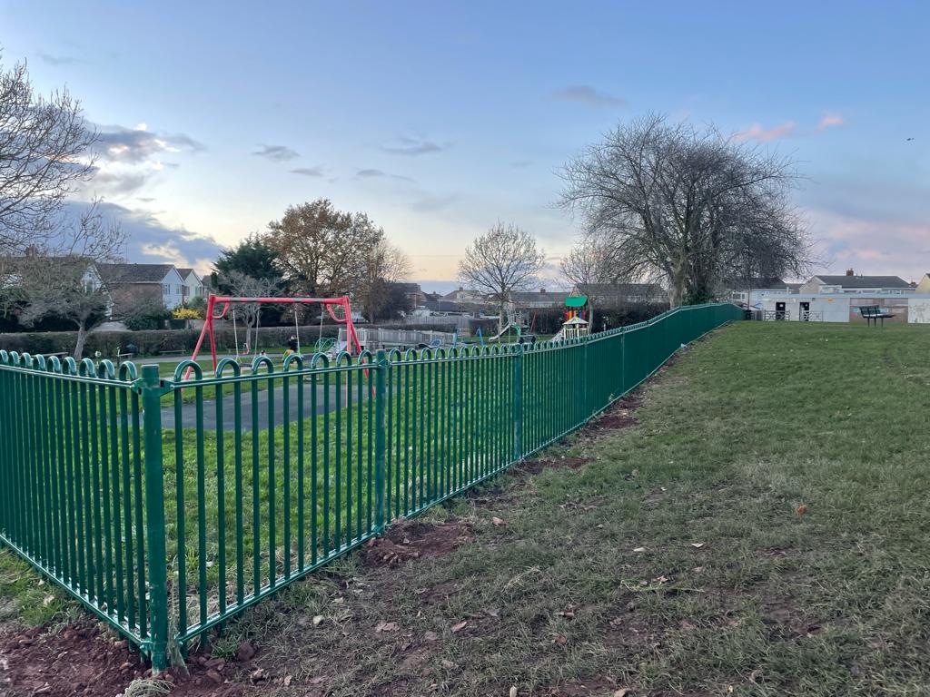 A park with a green metal fence, hinting at expert fencing installation, encloses a red playground structure. The grass is brownish and the trees are bare, signaling winter's grasp. In the background, a few houses sit under a cloudy sky.