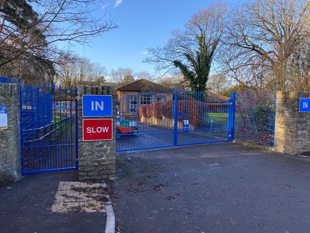 A blue metal gate with signs reading "IN" and "SLOW" blocks an entrance to a brick building, showcasing recent fencing installation. The area is paved with hard landscaping and lined with trees and bushes under a clear blue sky.
