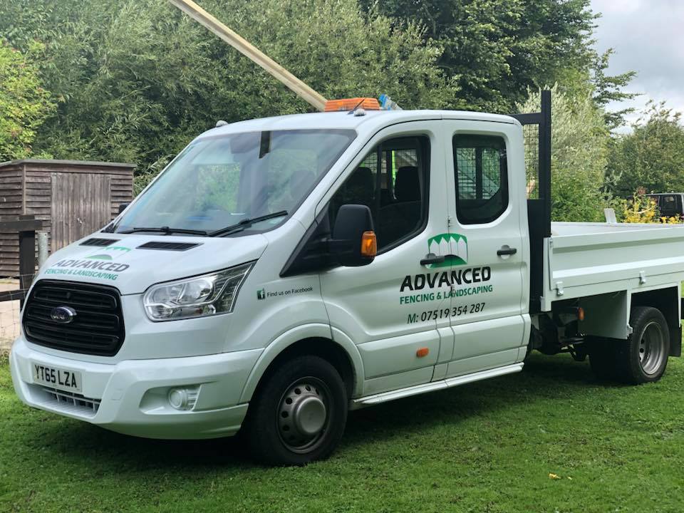 A white pickup truck featuring "Advanced Fencing & Landscaping" branding is parked on the grass. The open truck bed reveals tools for hard landscaping, while greenery and a wooden shed form the backdrop.