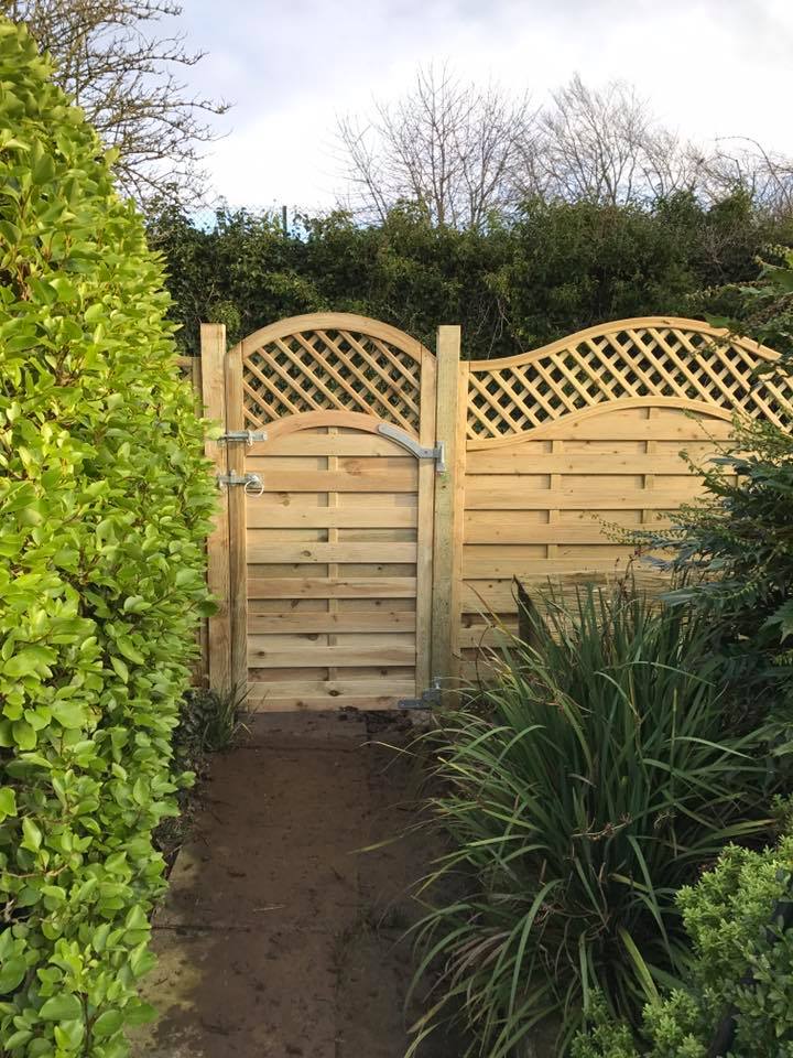 A wooden garden gate with an arched lattice top stands gracefully among lush hedges and plants. A narrow stone path, crafted with hard landscaping expertise, leads to this bespoke entrance. The overcast sky adds a serene atmosphere to the garden setting.
