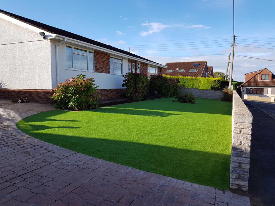 A house with a well-maintained front yard featuring a bright green artificial lawn is bordered by a brick wall and showcases bespoke railings. Shrubs near the house add charm, while a paved driveway lies to the left. New gates glisten under the clear, sunny sky.