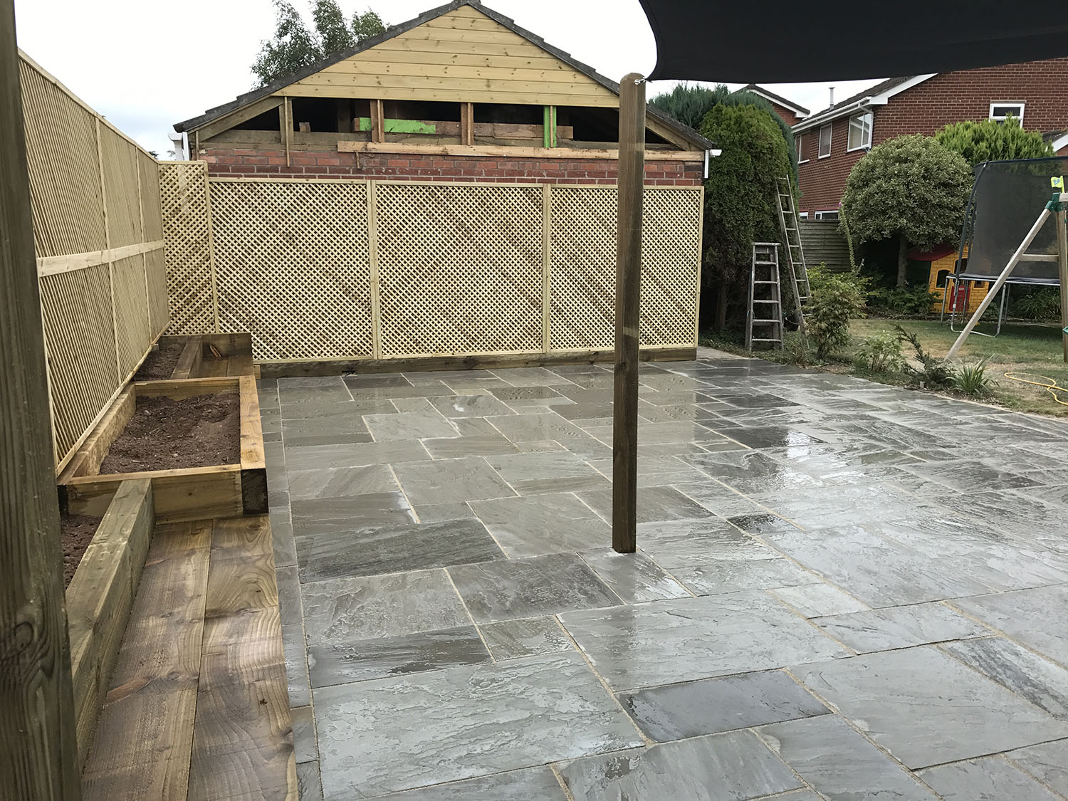 A newly paved patio features large gray stone tiles and a wooden planter on the left. A wooden post stands at the center, while lattice fencing partially covers a shed at the back. Thanks to expert fencing installation, a trampoline and ladder rest on the grass to the right, safely enclosed.