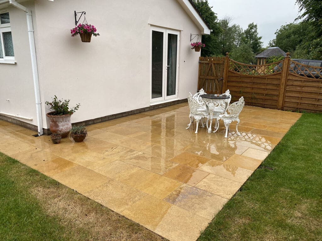 A patio with wet stone tiles sits next to a white house, featuring a white metal table and chairs. Potted plants line the wall, while hanging baskets bloom with pink flowers. A wooden fence, crafted by a fencing installation expert, encloses the area, with grass visible in the foreground.