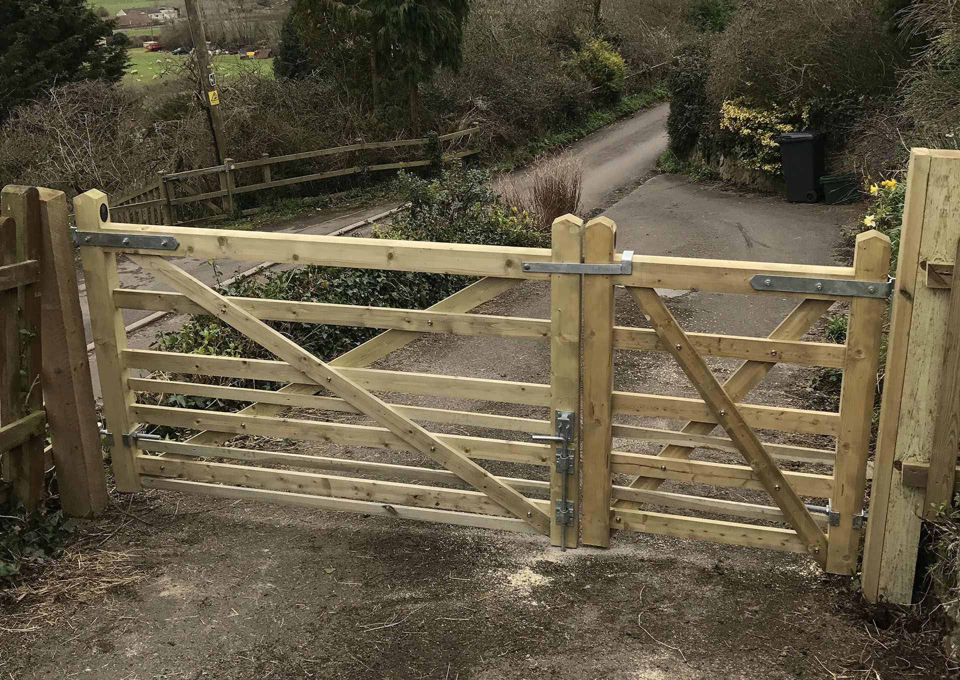 A wooden farm gate made of natural timber slats with a diagonal cross-brace stands snugly, showcasing bespoke railings. It's secured with metal hardware and opens onto a narrow rural road, surrounded by lush greenery and trees. A trash bin is visible in the distance.
