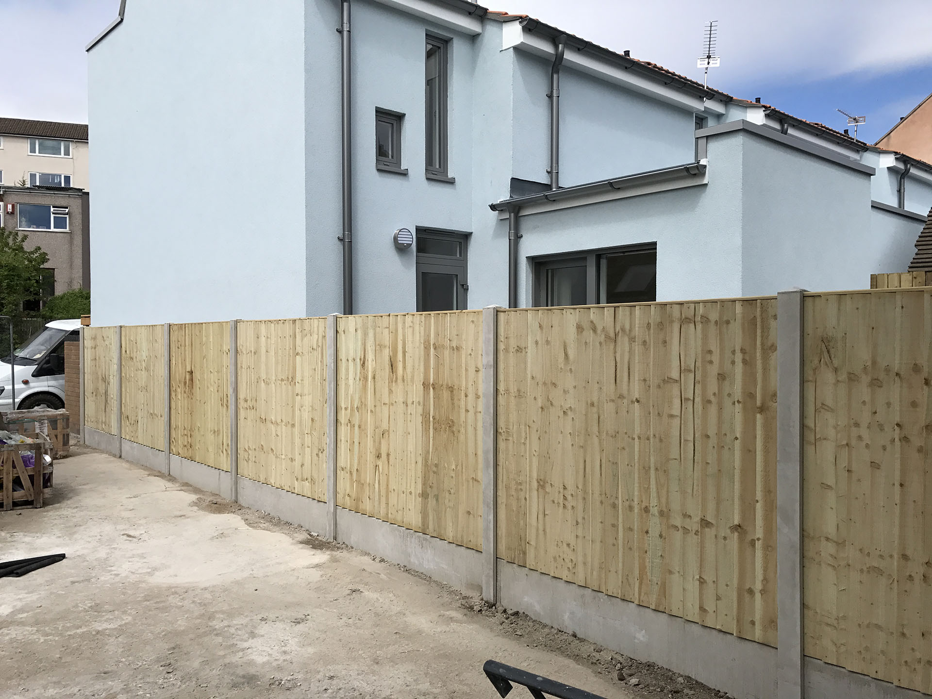 A modern two-story house with light blue walls and gray trim stands proudly, featuring a newly installed wooden fence by a renowned fencing installation team. A white van is partially visible in the driveway on the left, while the sky remains partly cloudy.