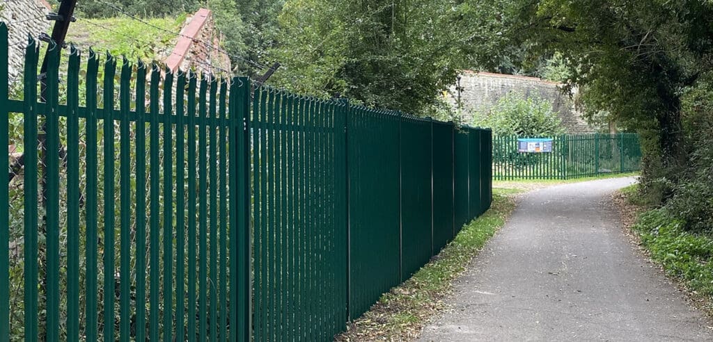 A paved path runs alongside a tall green metal fence with pointed tops, expertly installed by a commercial fencing contractor. Trees and dense greenery envelop the path, while a building with a slanted roof peeks through in the distance, near where new gates can be spotted by the sign.