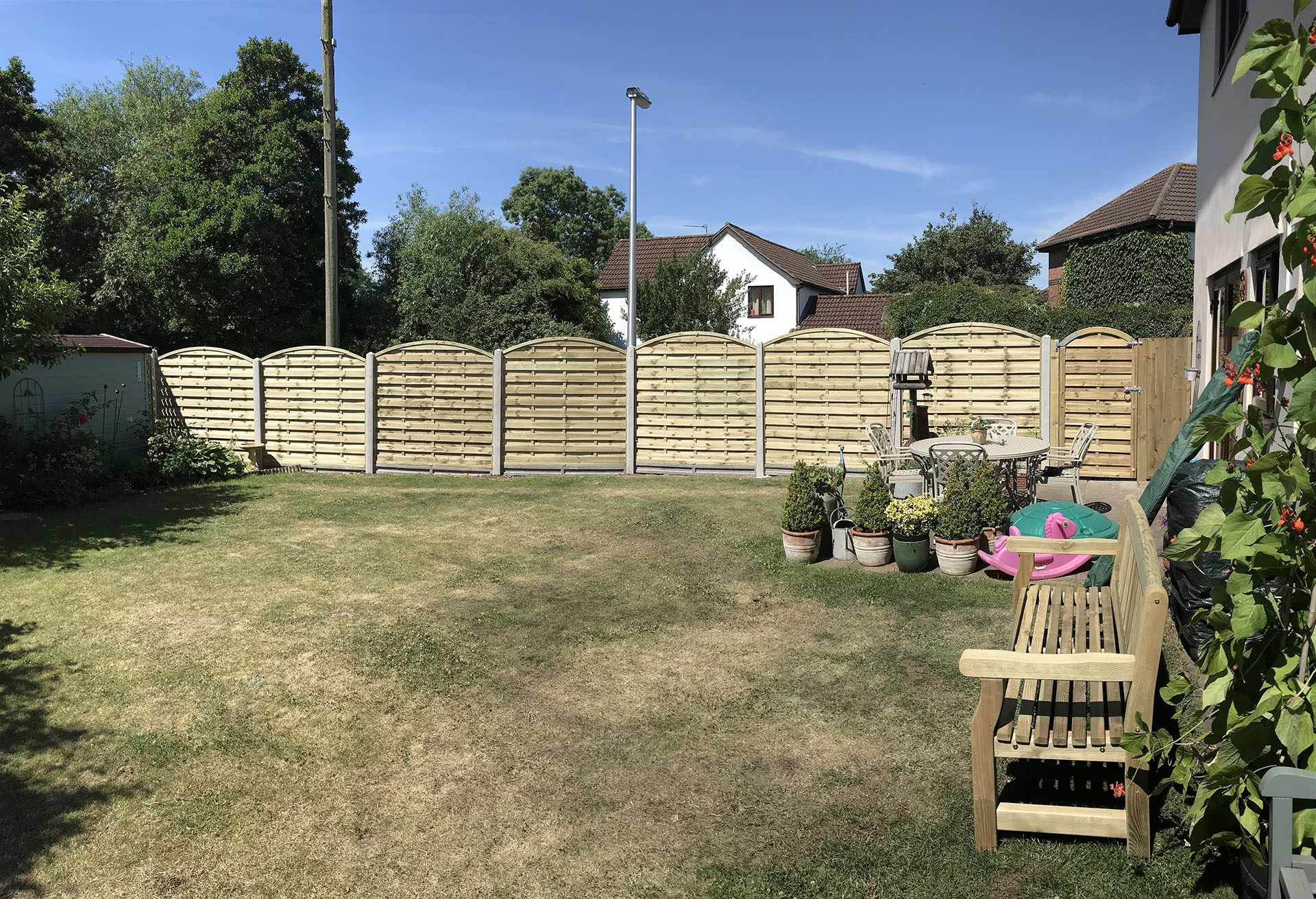 A backyard framed by a sturdy wooden fence, hinting at expert fencing installation. The green lawn has some dry patches, while potted plants rest near a wooden bench. A small table with chairs adds charm. Trees and houses are visible under the clear blue sky, creating a serene landscape.