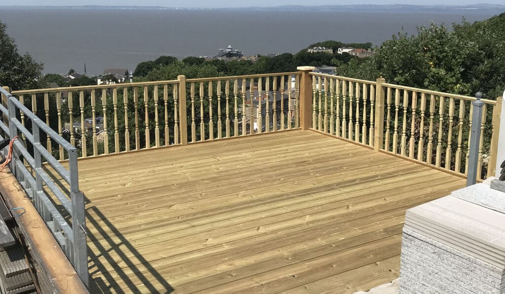 A wooden deck with new gates and railing overlooks a scenic view of the sea and distant hills. Elevated and surrounded by trees, it sits beneath a clear blue sky, with a few houses visible below the deck.
