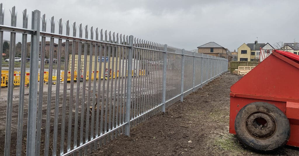 A long metal fence, installed by a commercial fencing contractor, stretches along the edge of a construction site. In the foreground, a large red skip is partially visible. Yellow barriers and containers are seen in the background, with cloudy skies overhead.