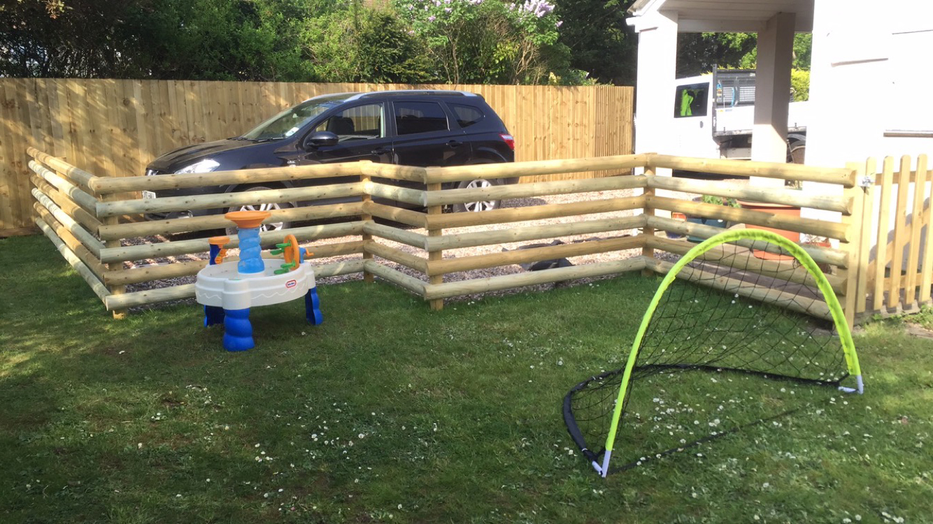 A backyard features a small black car within a fenced-off area, complemented by meticulous hard landscaping. The children's activity table sits by a small soccer goal on the grass. A new wooden fence lines the perimeter, with trees and the house standing gracefully in the backdrop.
