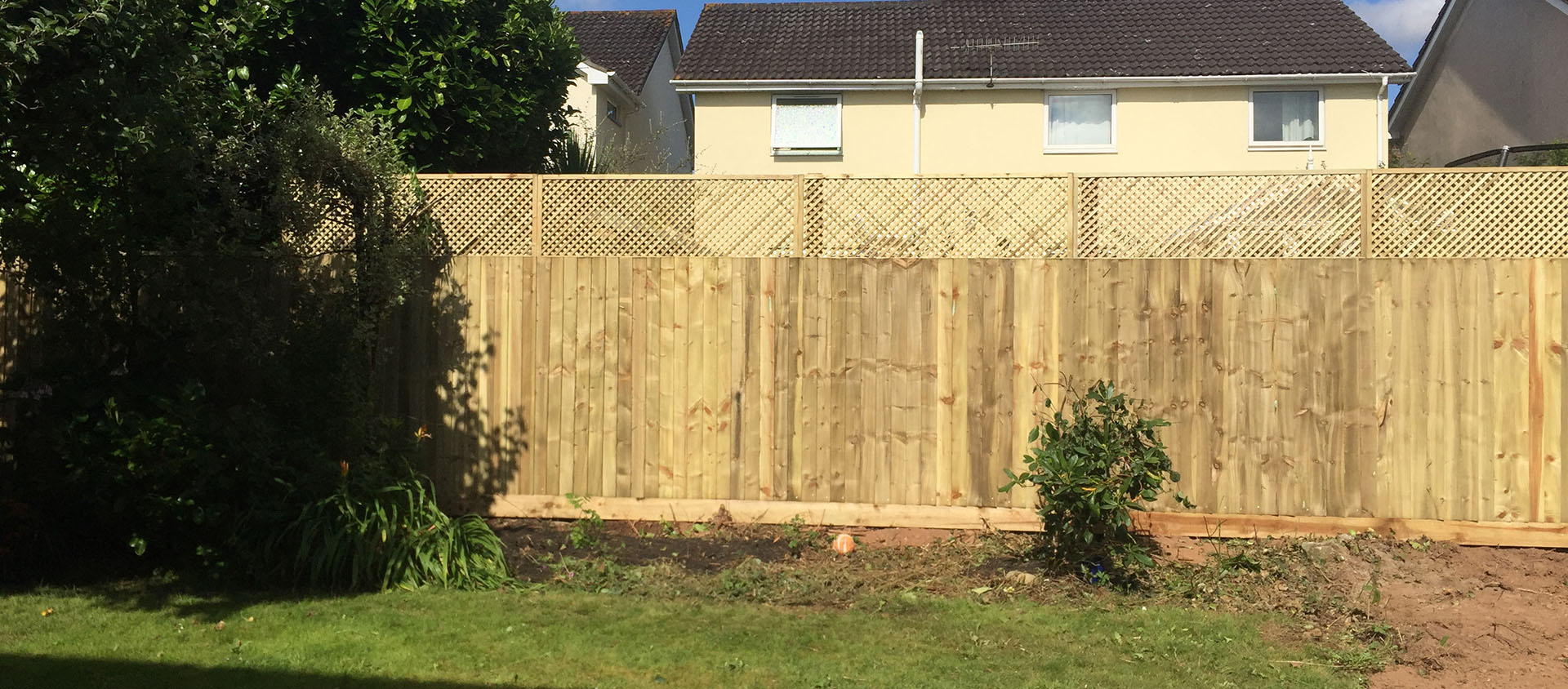A fenced backyard features a newly installed wooden fence with lattice detailing at the top, thanks to expert fencing installation. In front, small plants thrive. Two beige houses peek through under the bright blue sky, completing this picturesque scene.