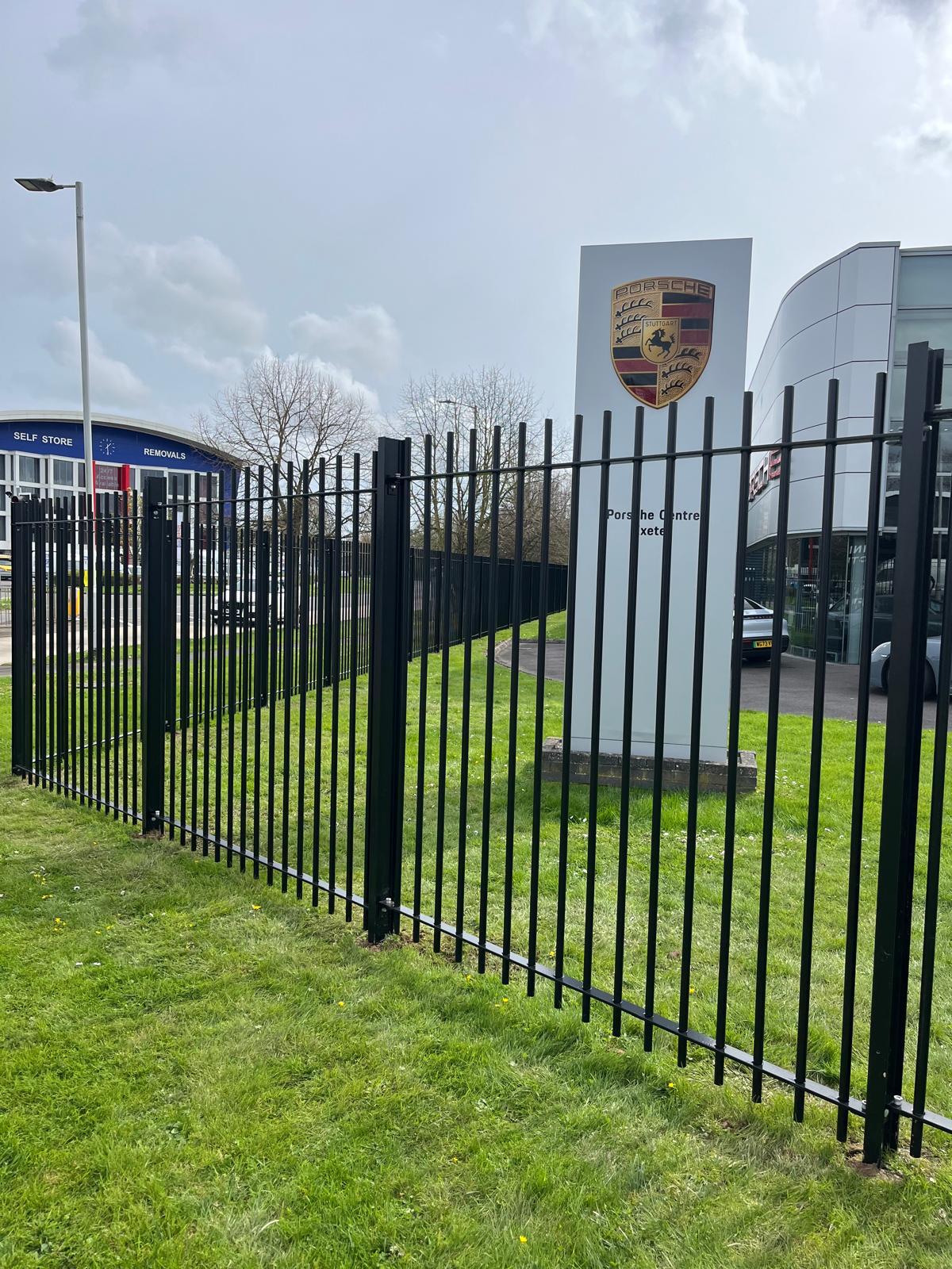 A car dealership features a tall white sign with the Porsche logo. A new black metal fence, the result of recent fencing installation and hard landscaping, runs along the grassy area in front. An overcast sky and nearby buildings provide a moody backdrop.