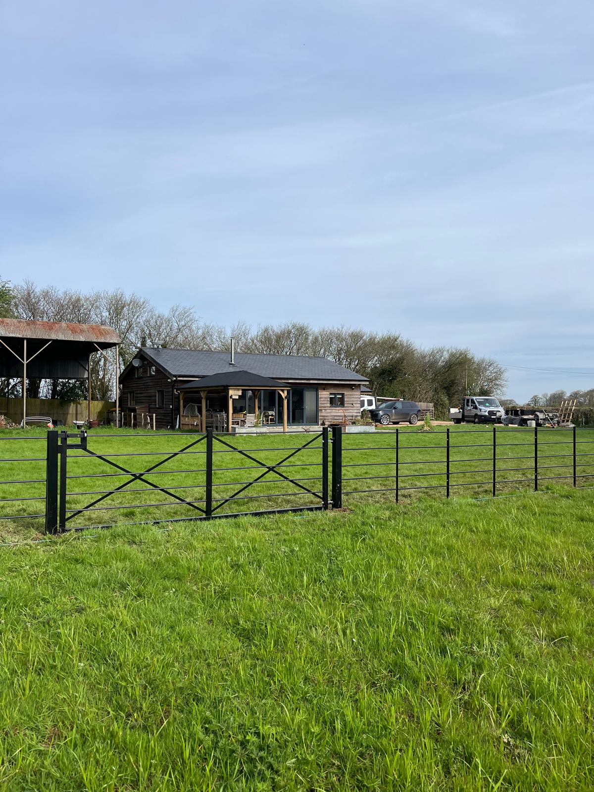 A rural scene reveals a small wooden house enclosed by metal fencing, perhaps the handiwork of a skilled fencing installation expert. Nearby, a truck and car rest quietly under a cloudy sky amidst trees. In the foreground, a grassy field stretches out serenely.