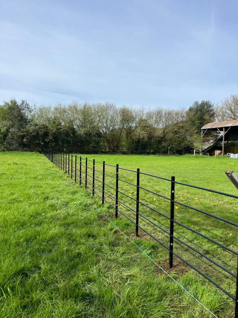 A black metal fence, crafted by a commercial fencing contractor, runs diagonally across the green grassy field under a clear blue sky. Trees form a boundary in the background, and a partially visible barn is on the right side.