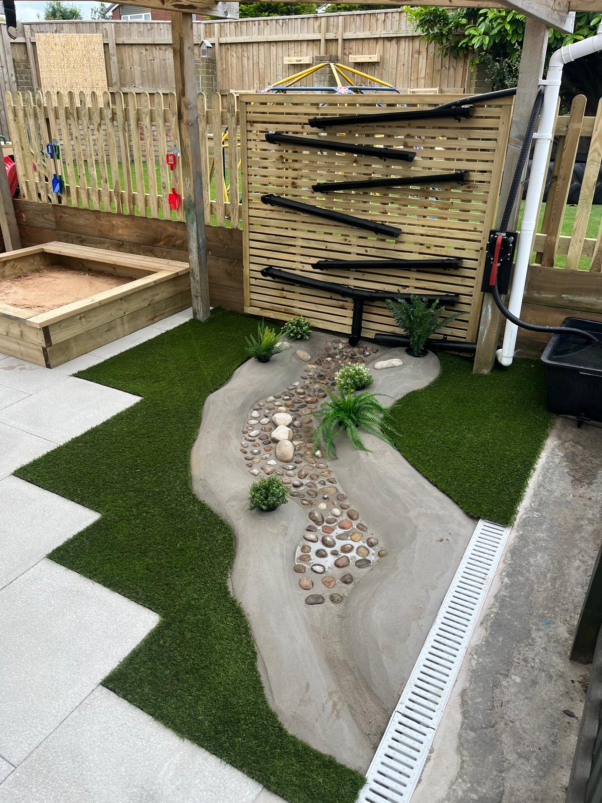 A children's outdoor play area features a sandbox, water play wall, and mini stream. The stream is lined with pebbles and artificial grass. Enclosed by professionally installed wooden fencing, the area includes a tiled floor with drainage for safety and cleanliness.