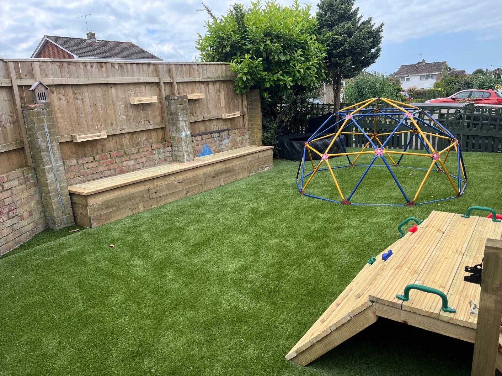 A backyard with artificial grass features a climbing frame with green handles, a geodesic dome play structure, and a wooden bench along the fence. New gates blend seamlessly into the backdrop of brick and wood, with a few houses visible beyond.