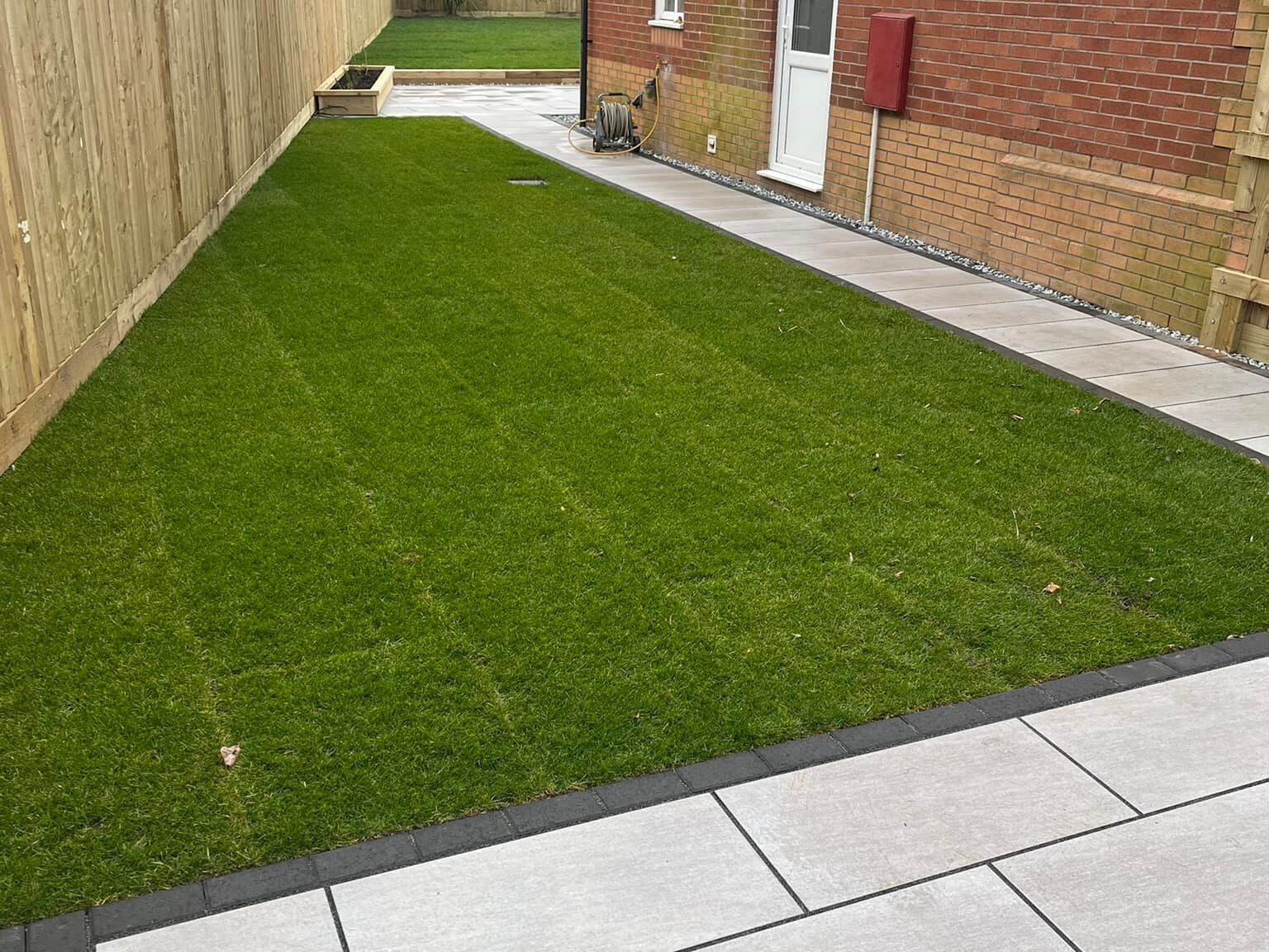 A backyard with a neatly manicured green lawn bordered by bespoke railings on one side and a brick house with a white door on the other. A paved patio area with large gray tiles is in the foreground. A small wooden planter is in the corner.